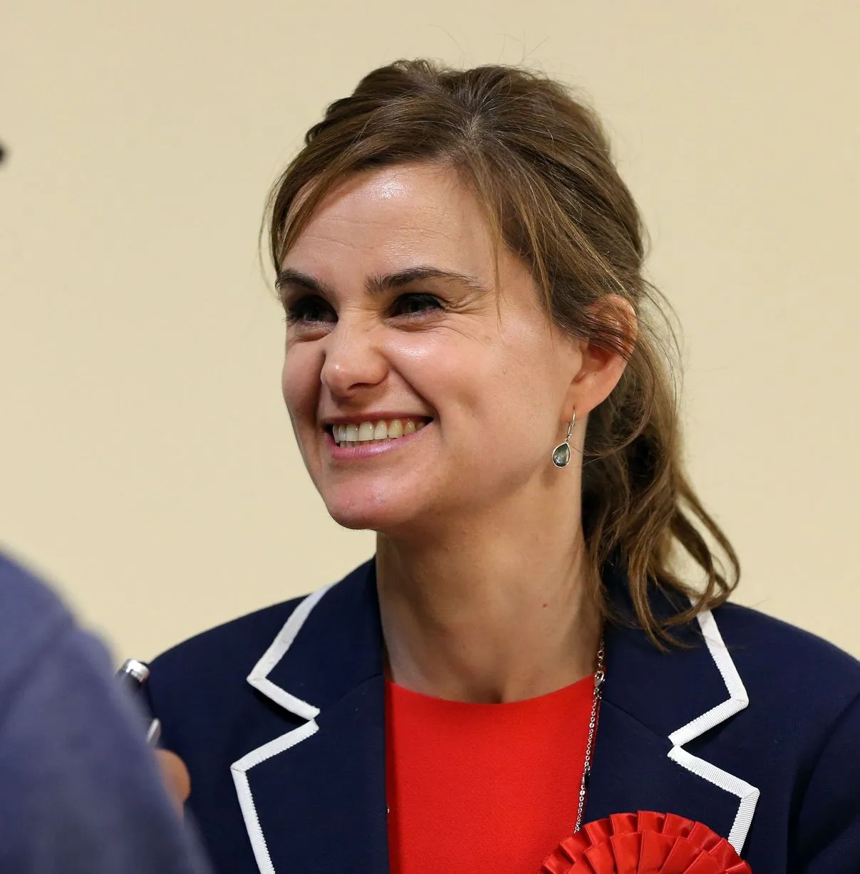 Jo Cox Image of Jo Cox smiling in a red top, wearing a navy blue blazer with white piping, and smiling.