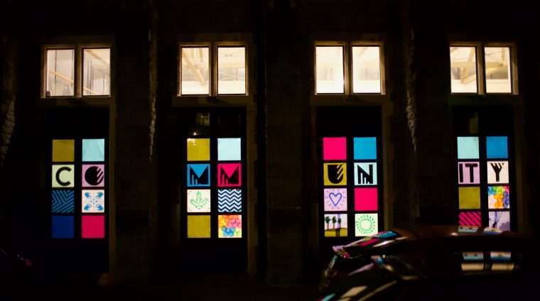 Picture of a building at night with the lights on inside. Four windows are decorated with coloured paper and the word 'Community'.