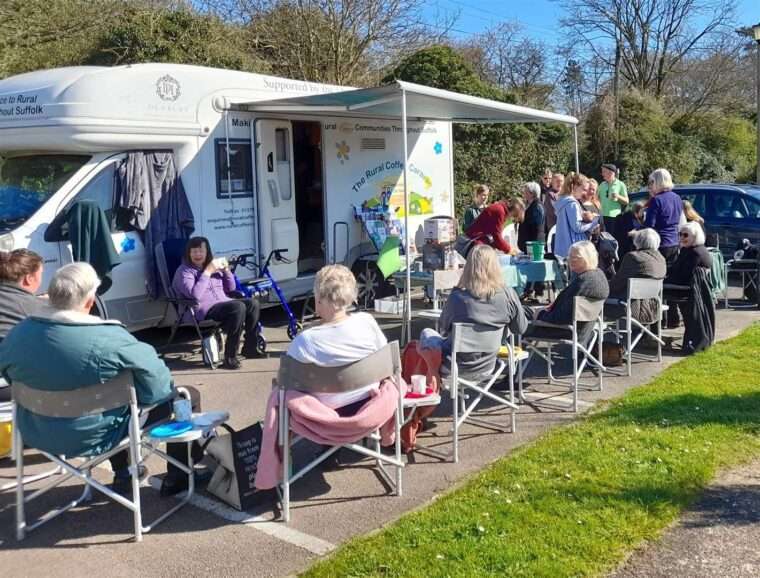 Photo shows the Rural Coffee Caravan with its door open and canopy up and people sitting around on camping chairs chatting whilst drinking coffee