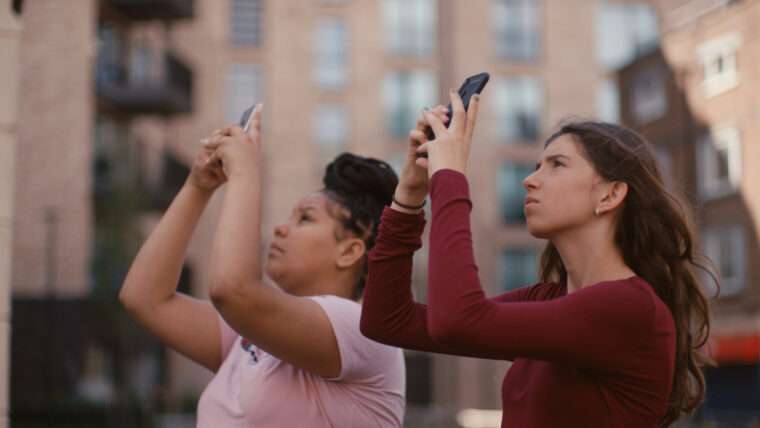 Two young women holding mobile phones aloft as they take a photo of something out of frame