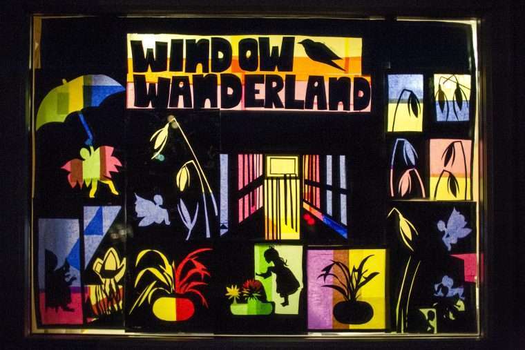 Photo of a window decorated for Window Wanderland. It has a piece of black paper which is cut out with sillouetts of flowers and fairies and the words Window Wanderland. THe holes in the paper are filled with coloured tissue paper and it's illuminated from behind to give a stained glass window effect.